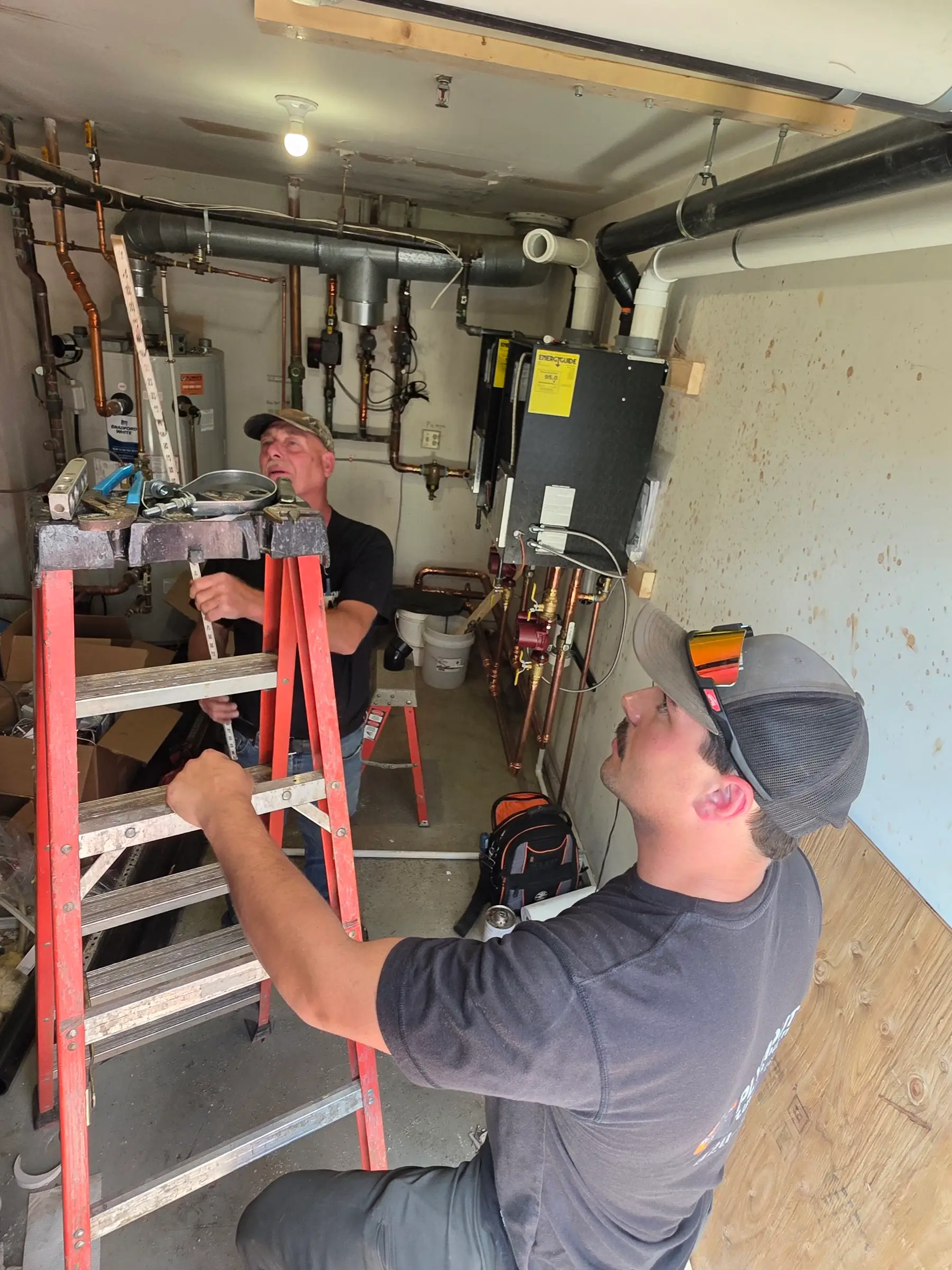 Two Plymouth Plumbing & Heating plumbers at work in a mechanical room beside a Bradford White water heater